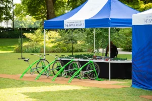A blue and white striped marquee stands on a lawn next to a row of stationary green bicycles used for an outdoor activity.
