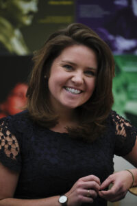 A woman with shoulder-length brunette hair and a bright smile stands in front of a coloUrful, blurred background. She is wearing a black lace-detailed top, a watch, and a ring while looking directly at the camera.