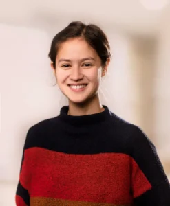 A young woman with dark hair pulled back smiles warmly at the camera. She is wearing a navy blue and red colour blocked jumper against a soft, out-of-focus neutral background.