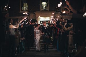 Newlywed couple walking through a tunnel of guests holding sparklers outside a grand building at night.