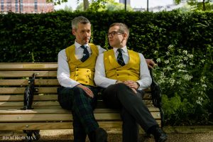 Two grooms in matching yellow waistcoats and ties sitting closely together on a park bench, surrounded by greenery.