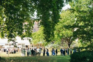 Wedding guests gathered in small groups on a sunny lawn outside a marquee, framed by large overhanging trees and lush greenery.
