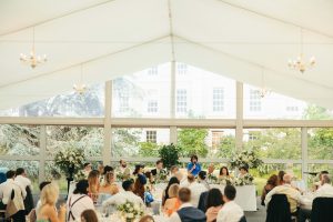 Wedding guests seated in a bright marquee, listening to a speech at the top table, with large windows framing views of trees and a historic building outside.