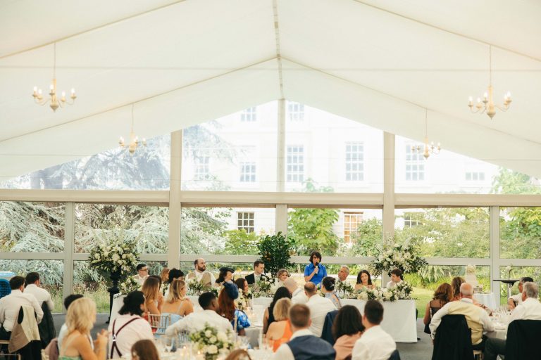 Wedding guests seated in a bright marquee, listening to a speech at the top table, with large windows framing views of trees and a historic building outside.