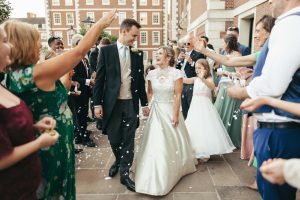 Bride and groom holding hands and smiling as guests throw confetti along a stone pathway outside a red brick venue.