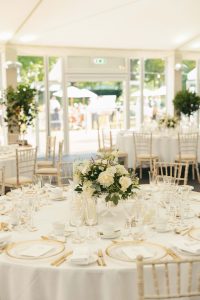 Round wedding breakfast table laid with white linen, gold-rimmed glassware and a central flower arrangement inside a bright marquee with garden views.