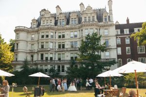 Wedding guests relaxing on lawn chairs and chatting under parasols in front of a large ornate white townhouse on a sunny day.