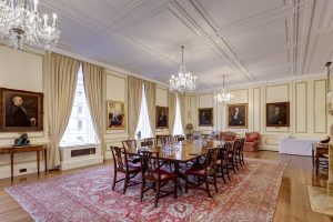 A long perspective of a formal dining or boardroom with heavy gold drapes, a large patterned rug, and rows of mahogany chairs.