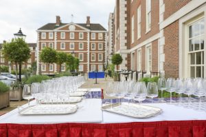 A wide shot of a terrace set up for an event, featuring multiple silver trays lined with wine glasses on white-clothed tables. The background showcases grand red-brick architecture with white window frames and a courtyard with spherical topiary trees.