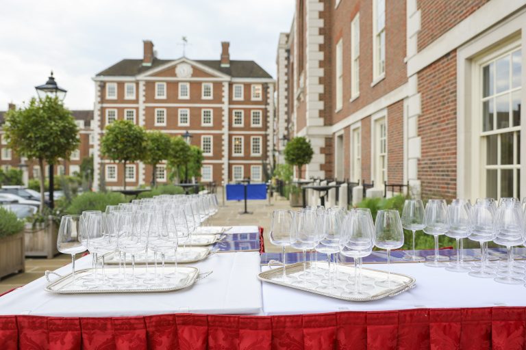 A wide shot of a terrace set up for an event, featuring multiple silver trays lined with wine glasses on white-clothed tables. The background showcases grand red-brick architecture with white window frames and a courtyard with spherical topiary trees.