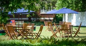 Wooden tables and chairs set up on a lawn under large blue umbrellas for outdoor dining.