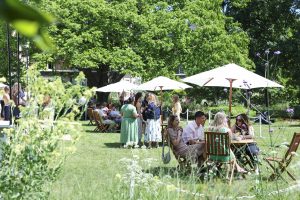 Guests relaxing and chatting at an outdoor summer event, seated at wooden tables under white parasols on a sunny lawn surrounded by trees and flowers.