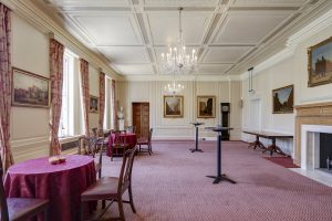 An airy reception hall with high coffered ceilings, small round tables with red linens, and tall cocktail tables on a pink patterned carpet.