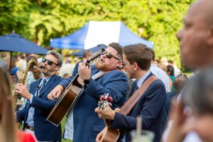 Live band performing at an outdoor corporate event, with musicians in suits playing guitar and saxophone in front of a crowd on a sunny day.