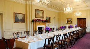 A long formal dining table set with white linens, purple floral centerpieces, and rows of wooden chairs.