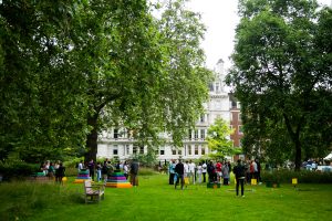 Wide garden lawn in front of a large white multi-story building with many windows, surrounded by tall green trees; group of adults chatting casually near colourful stacked chairs (pink, yellow, blue, green), croquet mallets and balls, and a wooden bench.
