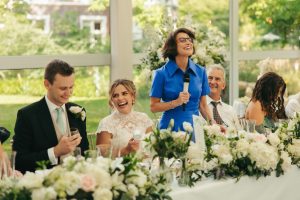 Wedding reception head table scene. A woman in a bright blue dress stands holding a microphone to give a speech, surrounded by seated guests, lush white floral arrangements, and large windows looking onto a green garden.