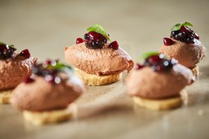 Close up of elegant bite-sized canapés, each with a smooth quenelle of pink mousse set on a round biscuit base, topped with glossy dark berry compote and a small green leaf garnish, with the sharply focused central piece surrounded by softly blurred matching canapés on a neutral background.