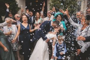 Joyful crowd of guests in elegant semi formal attire (suits, dresses, hats) cheering and throwing white confetti around a bride in white lace gown and groom in navy suit sharing a kiss on steps outside a grand white stone entrance