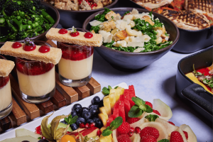 Close up of buffet table featuring small glasses of vanilla and raspberry dessert topped with shortbread, alongside a colourful fresh fruit platter and bowls of green salad